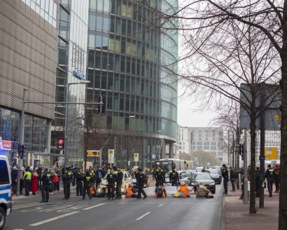 6 Klimaschützer protestieren auf der Straße sitzend. Einer der Klimaschützer, Christian Bläul hat eine beige Jacke an und blockiert einen Audi. Um sie herum stehen viele Polizist:innen.